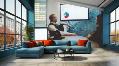 A business meeting in a boardroom with a projection: two caucasian partners in a defocused foreground looking at plasma screen with charts while sitting at a round table in an office with a laptop Wall mural