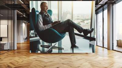 A cheerful adult successful man entrepreneur having a video group call with his partners via the laptop while sitting on a teal armchair in front of an office window with an urban cityscape outside Wall mural