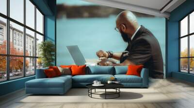 A dapper bald bearded black guy with glasses is looking at his watch. A handsome businessman is waiting for a person late for a meeting at a coffee shop. An elegant man alone at an outdoor table Wall mural