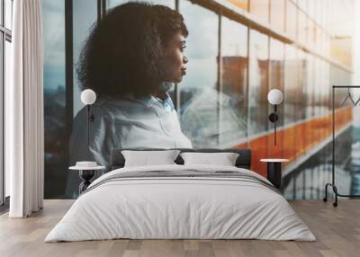 A dazzling young African-American woman entrepreneur with a cellphone in her hands is standing next to a panoramic window of a modern office skyscraper with orange lines and pensively looking outside Wall mural