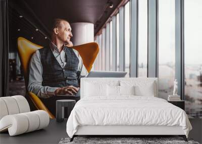 A handsome cheerful caucasian man entrepreneur is sitting with a laptop on his knees on an orange armchair next to the window indoors of a luxurious interior of a modern business office skyscraper Wall mural