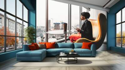 A ravishing African-American businesswoman on a yellow armchair is using her tablet PC; biracial woman entrepreneur using a digital tablet being on the top floor of a luxury business office skyscraper Wall mural