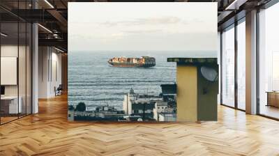 A telephoto shot of a large container ship sails on the open ocean near the coast. The vessel is loaded with multiple colorful cargo containers captured from a vantage point overlooking a coastal city Wall mural