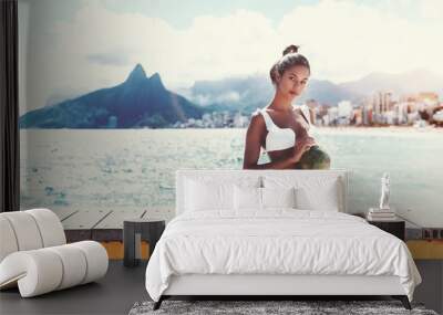 Charming young Brazilian girl is sitting on wooden bench next to beach and holding green coconut for drinking with cityscape of Rio de Janeiro and the Two Brothers hills in blurry background Wall mural