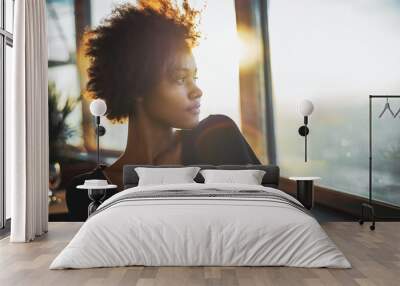 Curly charming young afro american tourist female in black dress is sitting in luxury restaurant of cruise ship near window and thoughtfully looking out the big window on ocean and warm sunset horizon Wall mural