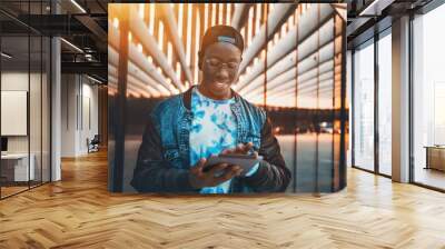 Handsome young black male student with white smile in glasses is checking his incoming messages via tablet pc; young handsome African guy waiting is chatting using digital pad near modern glass facade Wall mural