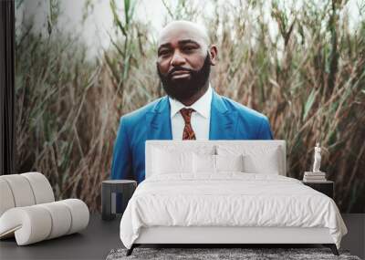 Portrait of a handsome mature bearded bald African man standing in front of thickets of reeds in an elegant blue business costume with a tie; a fancy serious black guy in a formal suit outdoors Wall mural