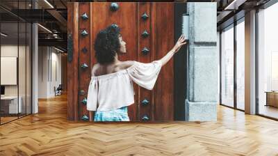 Side view of a dazzling African-American girl with a curly afro hair that is pushing a button of the house intercom outdoors in front of a huge wooden antique door with steel forged patches Wall mural