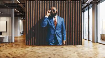 The portrait of a burly handsome bald black guy with a well-trimmed beard, in an elegant custom blue formal suit with a necktie, adjusting sunglasses outdoors in front of a wooden striped background Wall mural