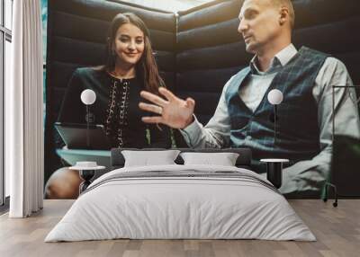 Two business colleagues are talking animatedly while sitting on an office sofa: a smiling businesswoman is listening to her colleague with a laptop that is actively gesturing in a defocused foreground Wall mural