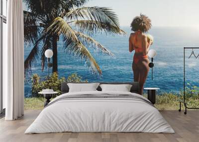 View from behind of a dazzling sexy African-American female in swimsuit enjoying the sun while standing on a resort shore next to a beautiful spreading palm and drinking coco water from a real coconut Wall mural