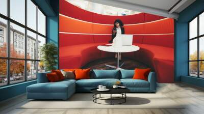 Wide-angle shot of a young charming African-American businesswoman sitting on a curved sofa of red color in an office lounge and using her phone; laptop, digital tablet, charging station on the table Wall mural