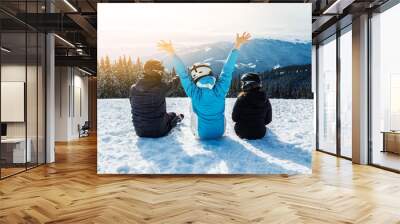 Three people in ski suits are sitting on the snow at the top of the mountain and looking into the distance. Wall mural