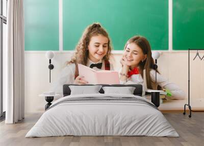 two brunette schoolgirls sit in the classroom at her desk with books on the background of the blackboard Wall mural