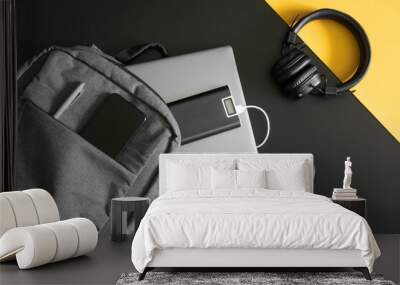 Gray textile urban backpack with laptop, external battery, phone, headphones, mouse and pen on black and yellow background. Travel, trip, study and lifestyle with gadgets. Recharging devices. Wall mural