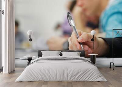 Guy student or schoolboy, writing a dictation, essay or filling out documents in the classroom, sitting at a desk next to other students. Photo. Selective focus. Close-up Wall mural