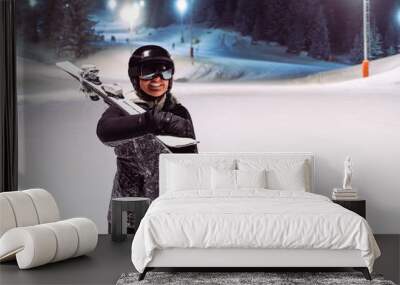 Happy woman skier with skis on her shoulder on a night ski slope Wall mural