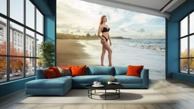 Slim girl in a black swimsuit is standing on a beach against the backdrop of a beautiful sunset. Wall mural