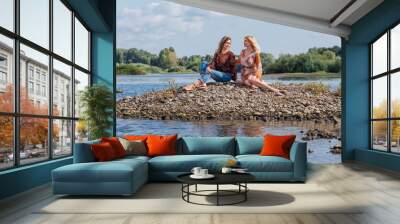 Two pretty sisters sitting on the small stone river island on a cloudy windy summer day, discuss girls secrets, laugh. Wall mural