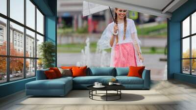 Young pretty girl with two braids in yellow boots and with transparent umbrella stands near fountain. Wall mural