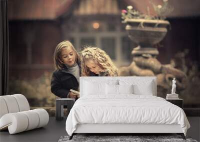 Cute long-haired blonde girl is reading a book sitting on the edge of old dried fountain in autumn. Funny boy is looking over her shoulder with funny expression. Image with selective focus and toning. Wall mural