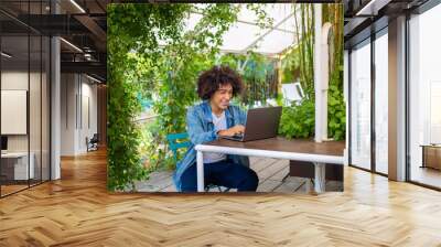 Smiling young mixed ethnic guy 20 years old, dressed in casual clothes, works on a laptop outdoors, in a green area in nature.Free Green open space for coworking, freelancer, relaxing, coffee, food.  Wall mural