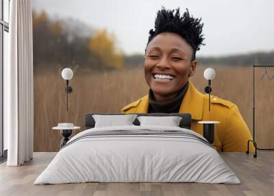 A joyful portrait of a woman smiling warmly outdoors in a natural autumn setting, wearing a yellow jacket that complements the earthy tones around her, capturing happiness and connection with nature. Wall mural