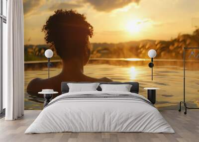 Silhouette of the back of an African American woman enjoying the sunset in an infinity pool, with a grass field on the horizon with the golden tones of the sunset  the water. Vacation and leisure Wall mural