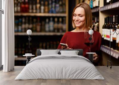 Image of happy blonde with phone and glass in hands at store with wine Wall mural