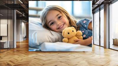 A girl child smiling, holding a soft toy and lying on a hospital bed in a modern hospital Wall mural