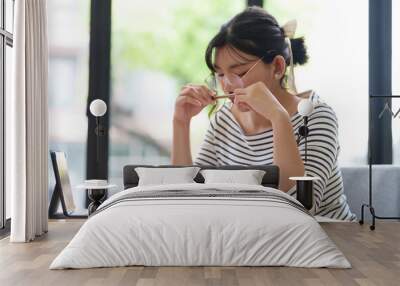 A young woman in a striped shirt is sitting at a desk, adjusting her glasses while reading a book. She is in a bright room with large windows, and appears concentrated on her task Wall mural