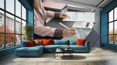 An Asian businesswoman writes on a digital tablet at her desk in a modern office. She is focused on her work, with documents and office supplies Wall mural