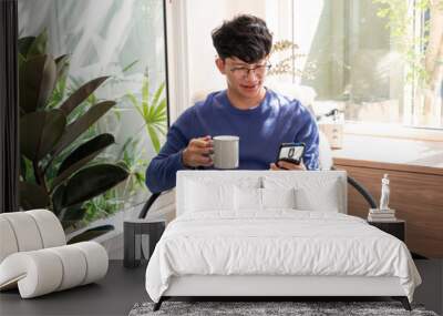 Handsome Asian teenager man wearing casual clothes in glasses using smart phone browsing Internet on mobile phone device at the living room. Wall mural