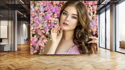 Brunette spring girl with long  and   shiny wavy hair .  Beautiful  model woman with curly hairstyle ,   background  wall of flowers . Skin Care, beauty and  spa Wall mural