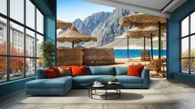 beach umbrellas and wooden lounge chairs on the sand of the beach against the backdrop of the sea and high rocky mountains in Egypt Wall mural