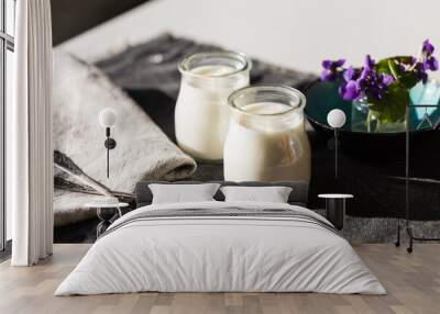 Two glass jars with natural homemade yogurt, spoons, little vase with violets on black fabric on white wooden table. Wall mural