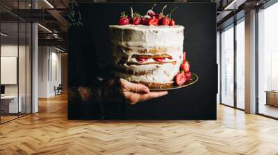 Woman in black embroidered dress holding strawberry and mascarpone layered Victoria sponge cake on dark background. Wall mural