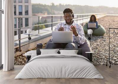 Adult African male wearing denim shirt and headset holding online conference over portable computer on rooftop, female colleague working on blurred background. Wall mural