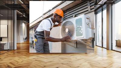 Adult male master wearing gray overalls and orange helmet leaning towards air conditioner and checking fan outdoor. African american man inspecting appliance while repairing in roof of building. Wall mural