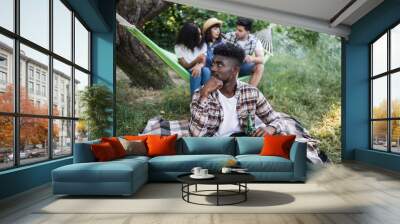 African american guy sitting on plaid with bottle of beer in hand and looking aside while his multiracial friends resting in hammock on background. Young people on picnic during summer time. Wall mural