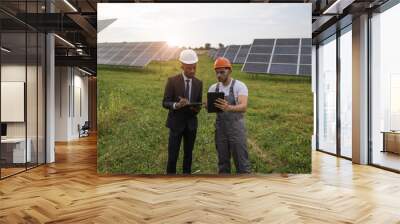 African american man in suit and indian man in uniform standing among solar plant and checking working process. People using digital tablet and clipboard during examination of panels. Wall mural