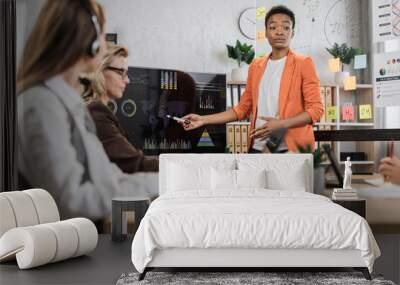 African american woman in orange suit showing financial statistic of enterprise during working meeting with diverse female colleagues. Cooperation, technology and people concept. Wall mural