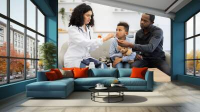 African female doctor offers medicine to young African boy with father at home. Scene shows caring healthcare professional and family in cozy setting emphasizing health child care and parental support Wall mural