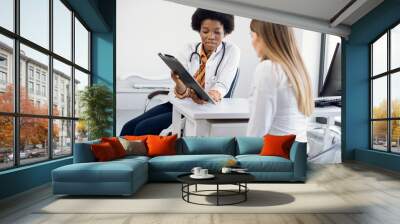 African female physician and young girl patient discussing something while sitting at the table. Medicine and health care concept. Doctor shows clipboard with information for patient Wall mural