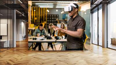 African young man wearing virtual reality glasses in modern interior design coworking studio. Smartphone using with VR goggles headset. Team of young people working at the table on the background Wall mural