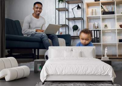 Afro american man sitting on couch and working on laptop while his little son playing with toys on carpet. Young father taking care of baby and working from home. Wall mural