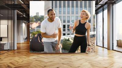 Athletic man in white t-shirt and woman in black top drinking water after jogging. Young couple having fun laughing after morning workout on the roof of the house. Wall mural