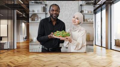 Attractive african american man and muslim young woman holding bowl with healthy salad with fresh vegetables while cooking dinner together and having fun in a new light kitchen. Wall mural