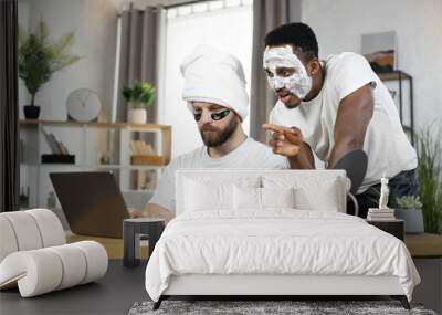 Attractive cheerful young multiracial two male friends with eye-patches and facial mask laughing when watching funny videos or photos on computer at home. Men skin care at home Wall mural