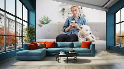 Attractive young woman in cozy outfit holding smartphone while her small pet having rest by her side on gray sofa. Active pet owner sharing online video of great outdoor fun with furry friend. Wall mural